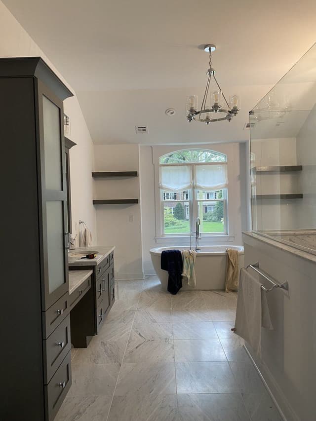 The primary bathroom of a Westfield, NJ new-construction home before renovation, with a freestanding tub under the window between two gray-stained cabinets