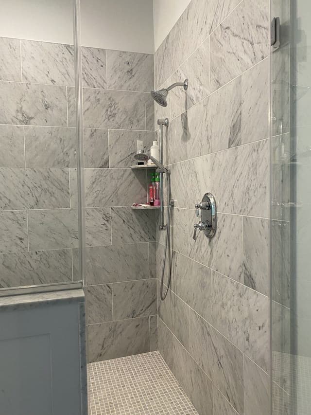 The shower of a Westfield, NJ new-construction home before renovation, lined in large-format marble-look tile behind a glass panel
