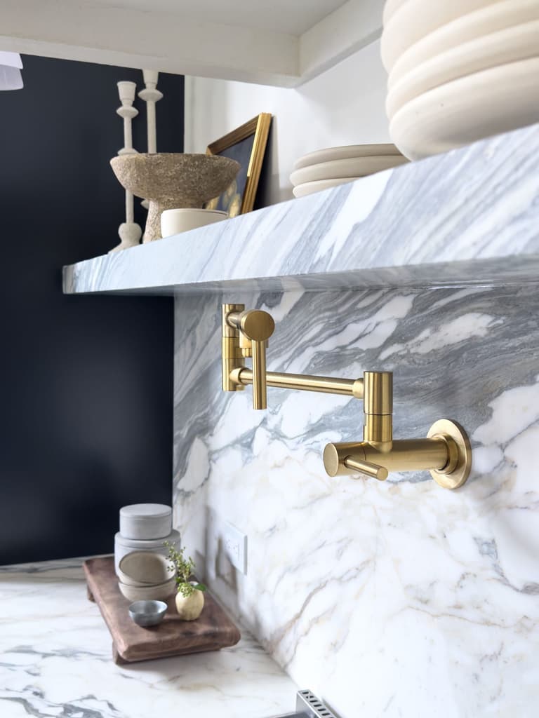 Unlacquered brass pot filler set through a marble slab backsplash below a floating marble shelf in a Mendham, NJ kitchen by Michael Robert Construction
