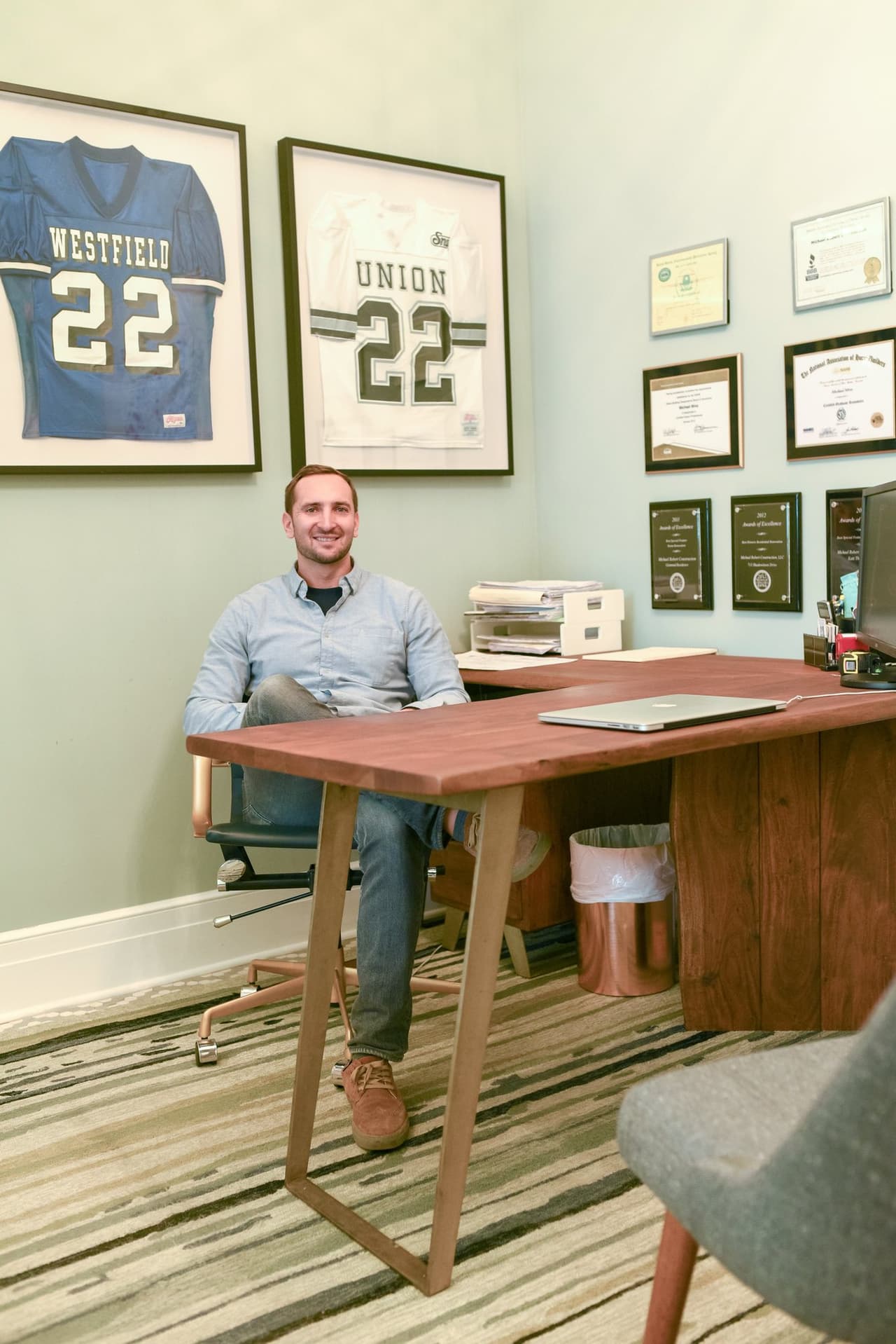 Mike Mroz at his walnut desk in the Michael Robert Construction office in Westfield, NJ, with framed Westfield and Union football jerseys on one wall and the firm's award plaques on the other