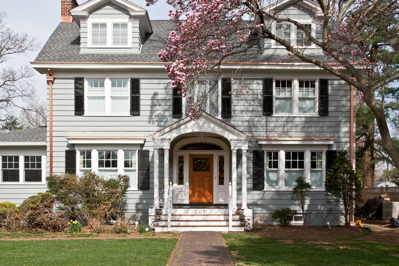 Front elevation of a gray shingled Colonial Revival with black shutters, an arched portico and a magnolia in bloom in Westfield, NJ