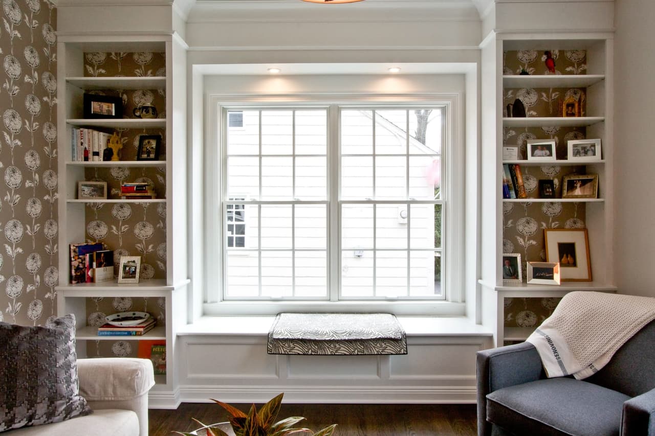 Window seat flanked by built-in shelving with patterned wallpaper behind, and an armchair alongside