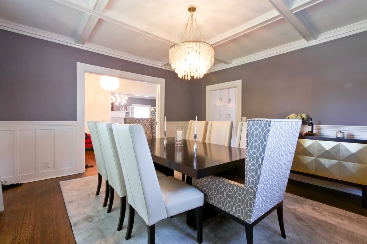 Dining room with a dark table, high-backed upholstered chairs, board wainscot and a crystal chandelier