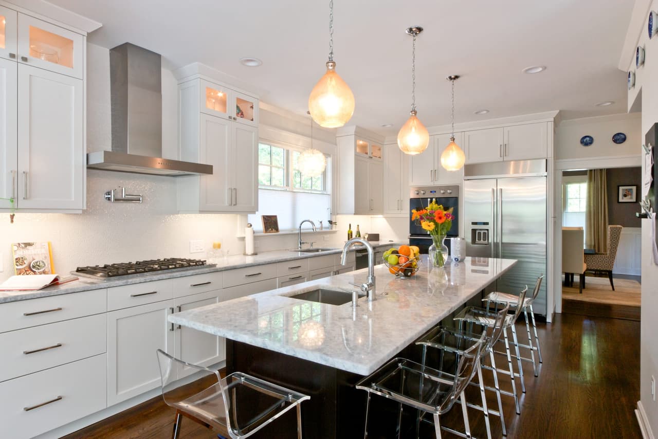 Kitchen with a stainless chimney hood over the range, marble counters and glass pendants above the island