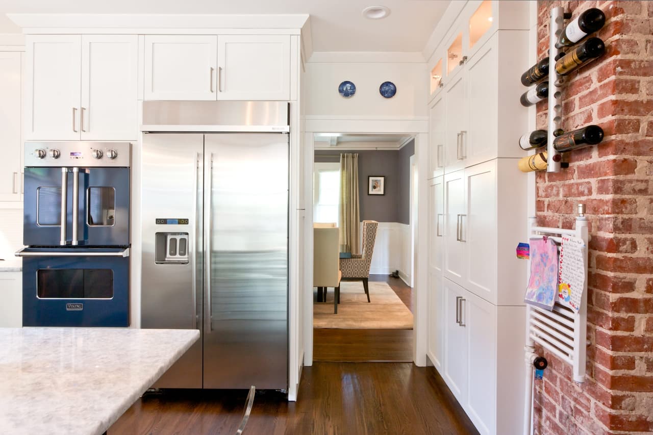 Kitchen of a Westfield, NJ renovation with a stainless refrigerator between white cabinets, blue wall ovens and a wine rack on the exposed brick chimney