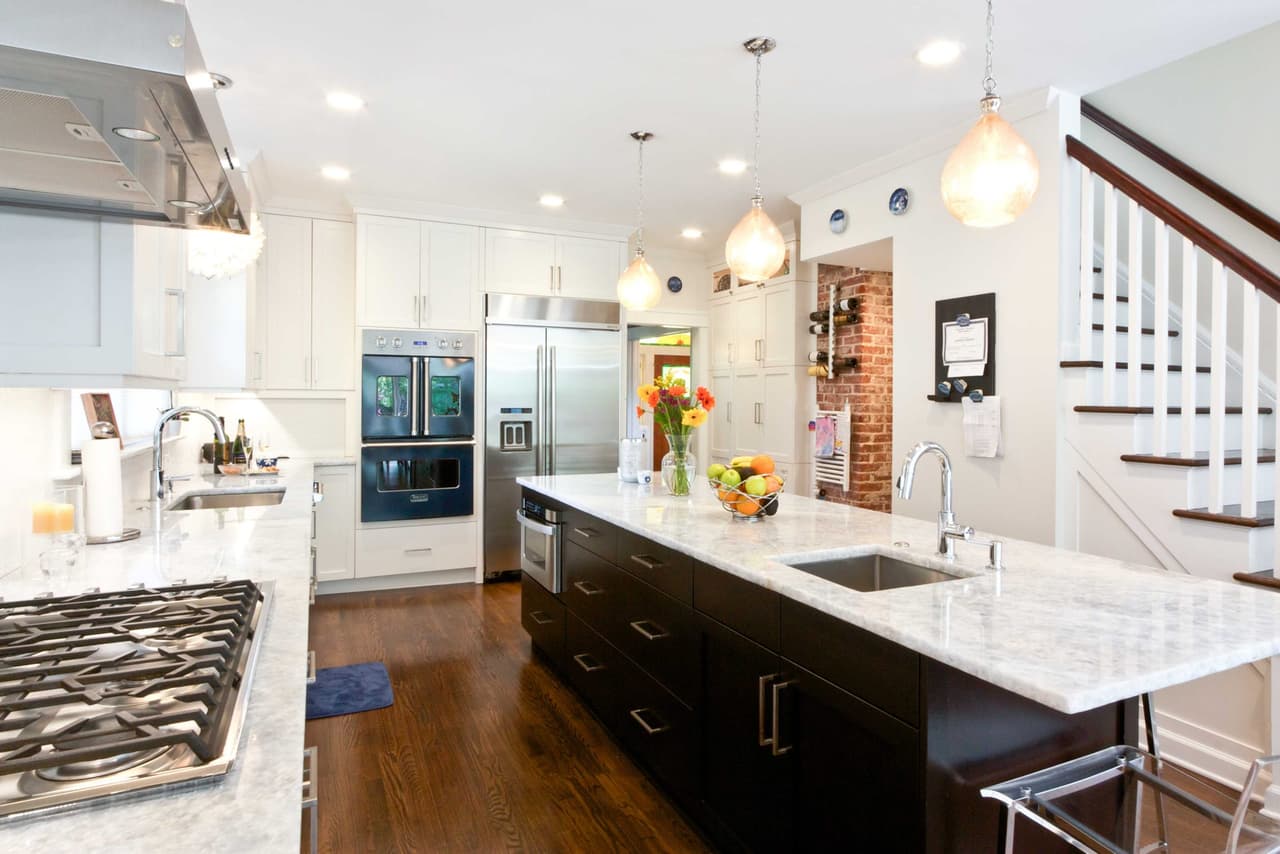 Kitchen with a long dark island, marble counters, glass pendants and a stair with a dark handrail beyond