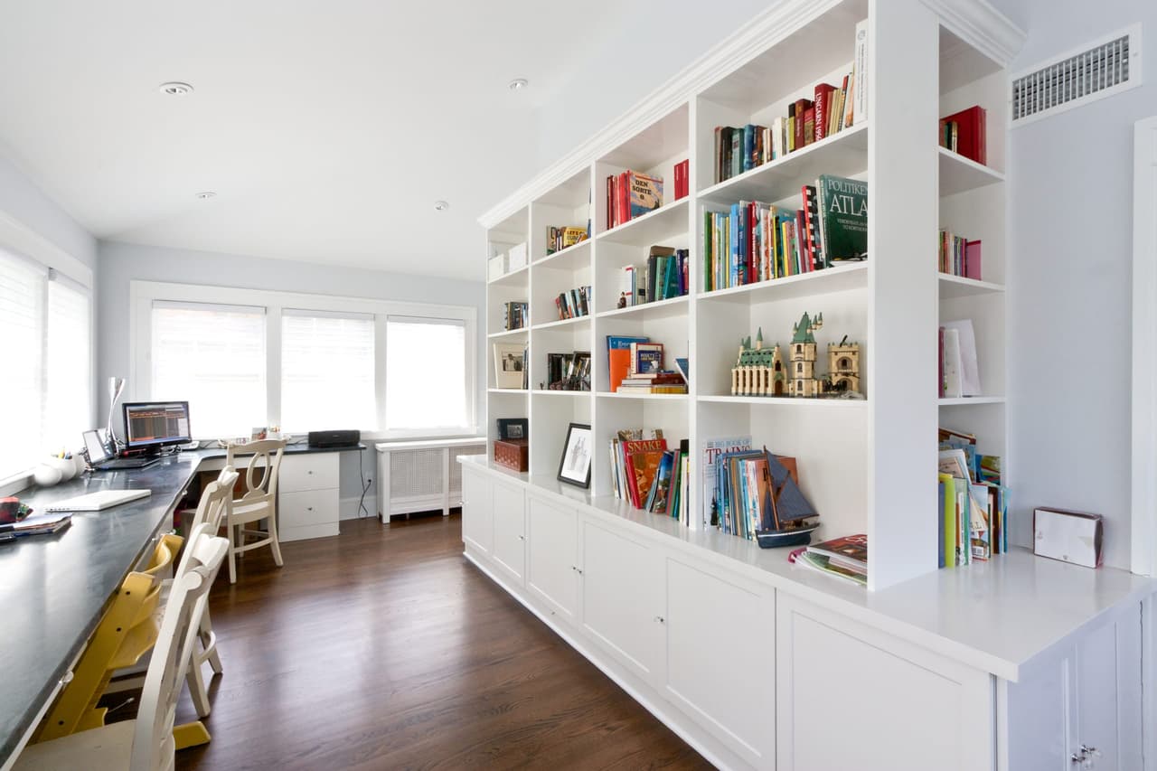 Built-in bookshelves and cupboards along the wall of a Westfield, NJ study, with the desk run and windows to the left