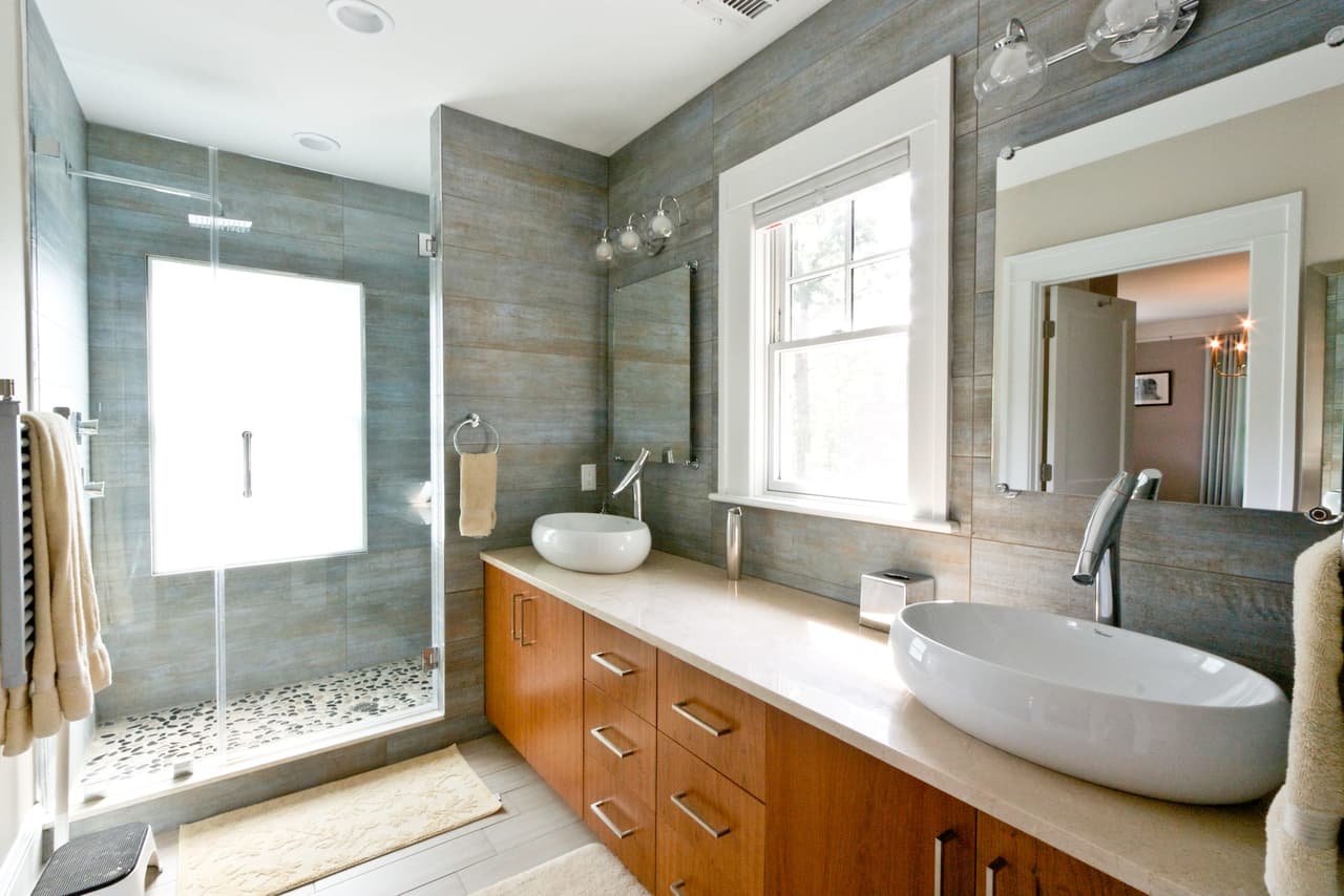 Bathroom in a Westfield, NJ renovation with twin vessel basins on a cherry vanity, a stone-tiled shower behind glass and a pebble floor