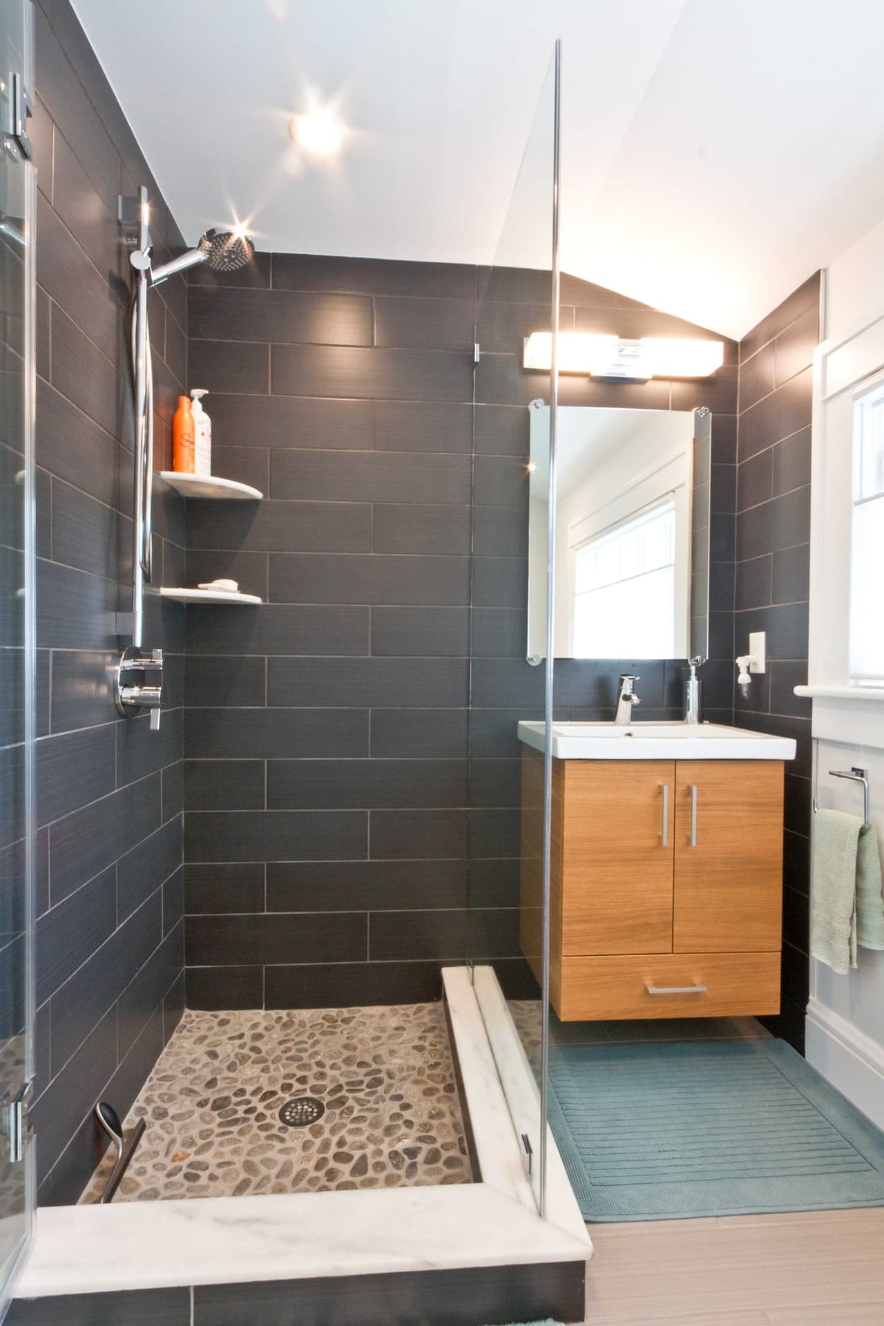 Shower with charcoal tile, a pebble floor, a glass partition and a wood vanity beyond
