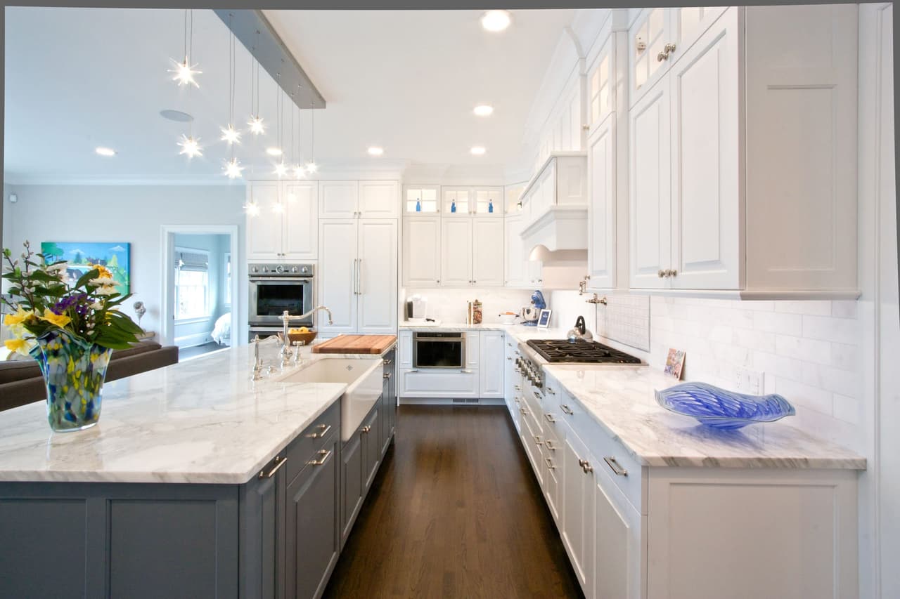 Kitchen with a long gray island, marble counters and a run of white cabinetry under a dropped beam