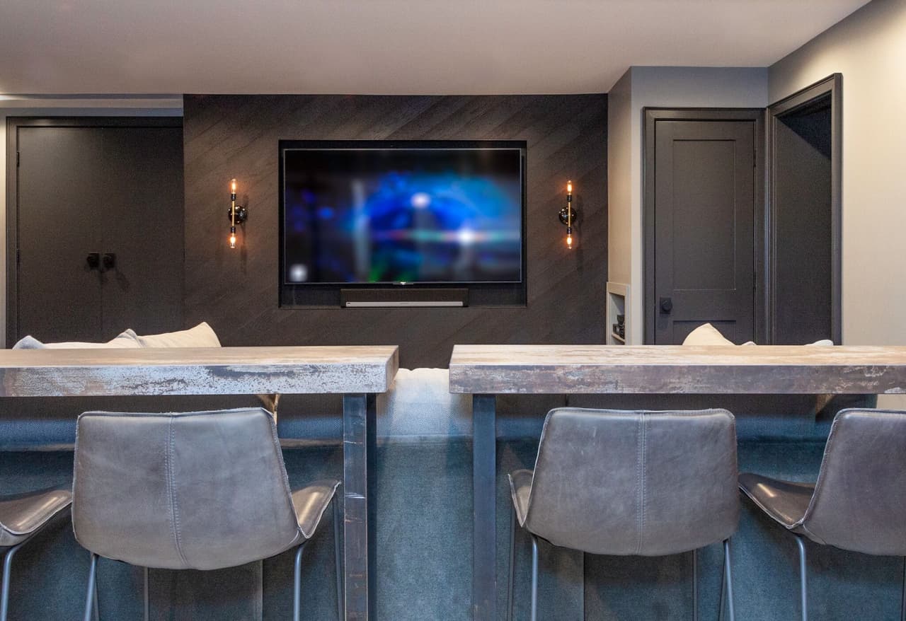 Lower-level bar with a dark paneled wall, a wall-mounted television, sconces and gray leather stools