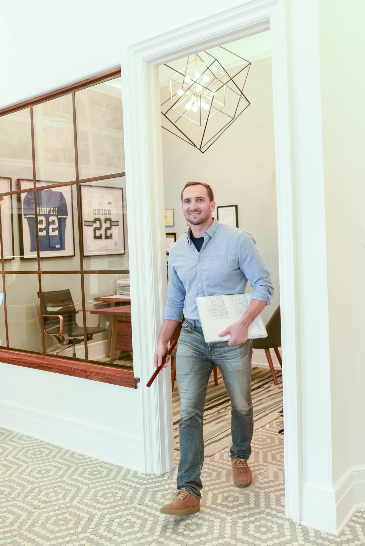 Mike Mroz of Michael Robert Construction in a cased doorway beside an interior steel-and-glass office window, carrying drawings