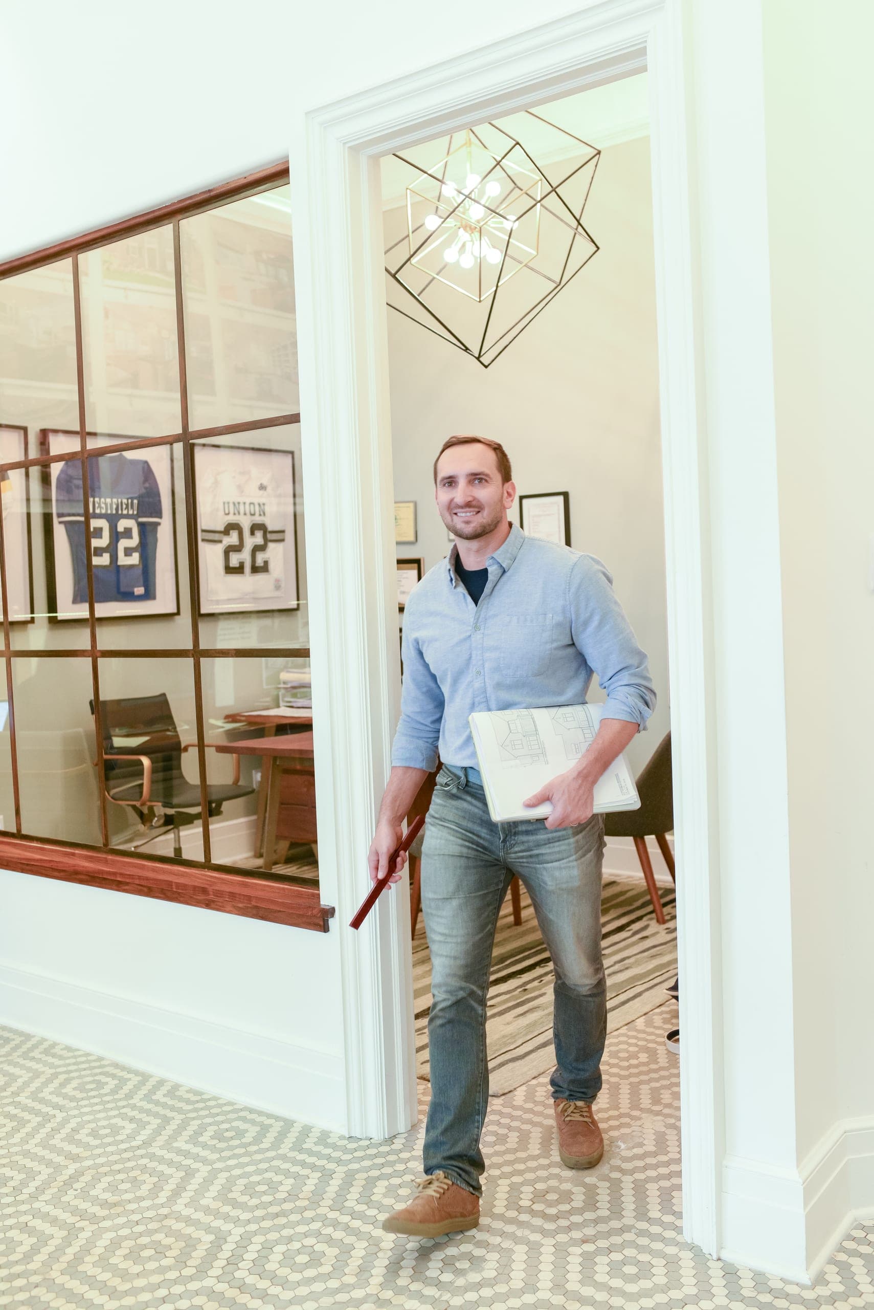 Mike Mroz of Michael Robert Construction in a cased doorway beside an interior steel-and-glass office window, carrying drawings