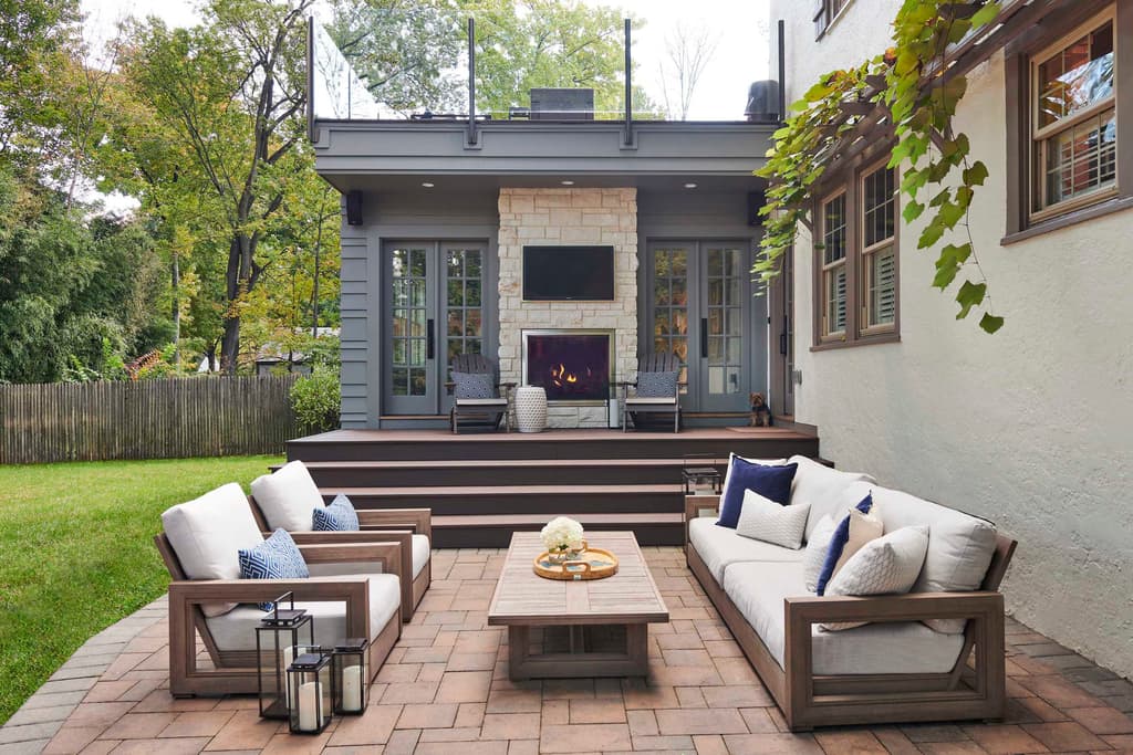 Covered outdoor living area at a Westfield, NJ home, with a stone fireplace and television under the porch, teak sofas on brick paving and lanterns on the steps
