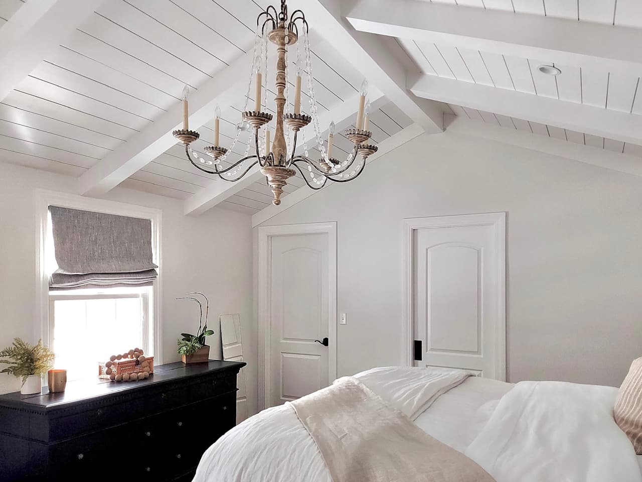 Primary bedroom of a Westfield, NJ renovation under a vaulted plank ceiling with exposed rafters, with an iron chandelier and a black dresser at the window