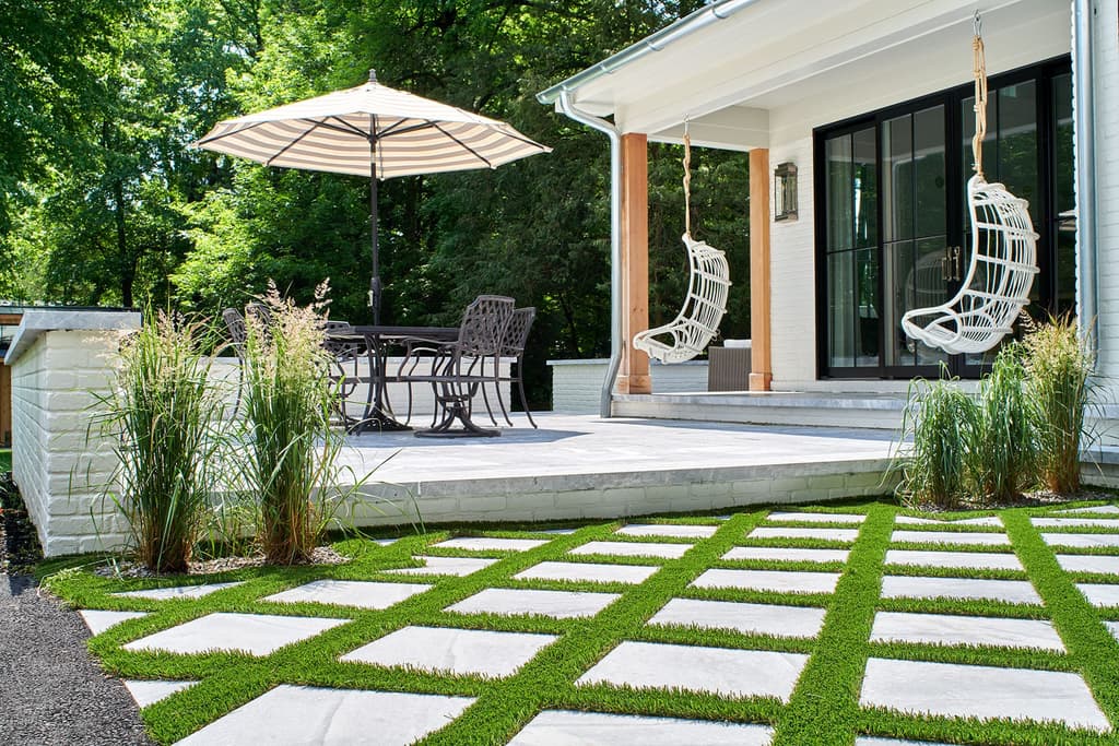 Raised stone terrace and white brick porch with hanging rattan chairs, above square pavers set in a grass grid