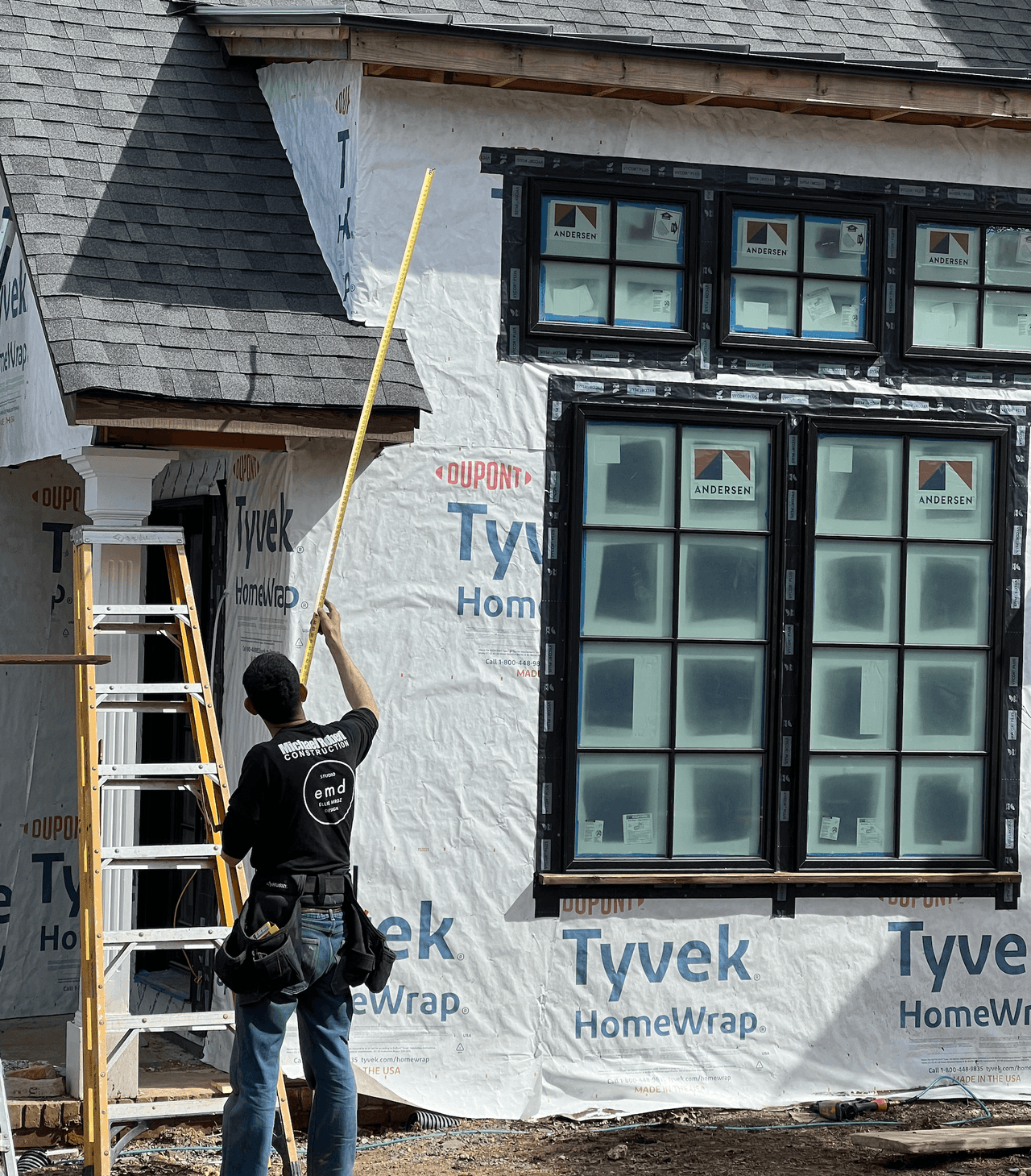 MRC carpenter on a ladder measuring a Tyvek-wrapped exterior wall with new Andersen windows installed, mid-construction
