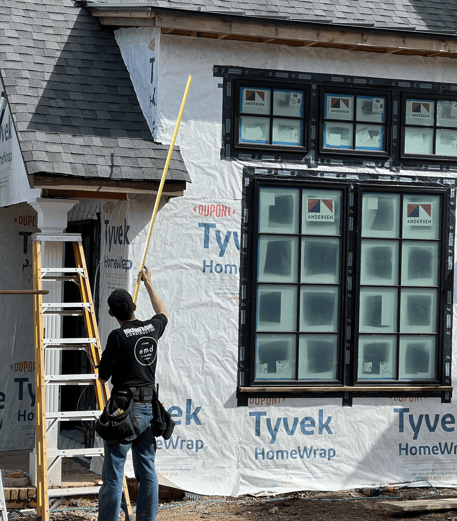 MRC carpenter on a ladder measuring a Tyvek-wrapped exterior wall with new Andersen windows installed, mid-construction