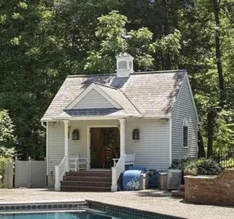 The poolhouse at a Mendham, NJ property before renovation, white clapboard with a cupola and a small porch above the pool