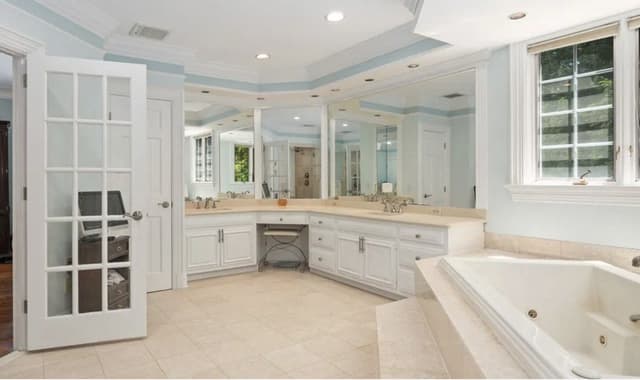 The primary bathroom of a Mendham, NJ house before renovation, with a corner jetted tub, a mirrored vanity run and pale blue walls