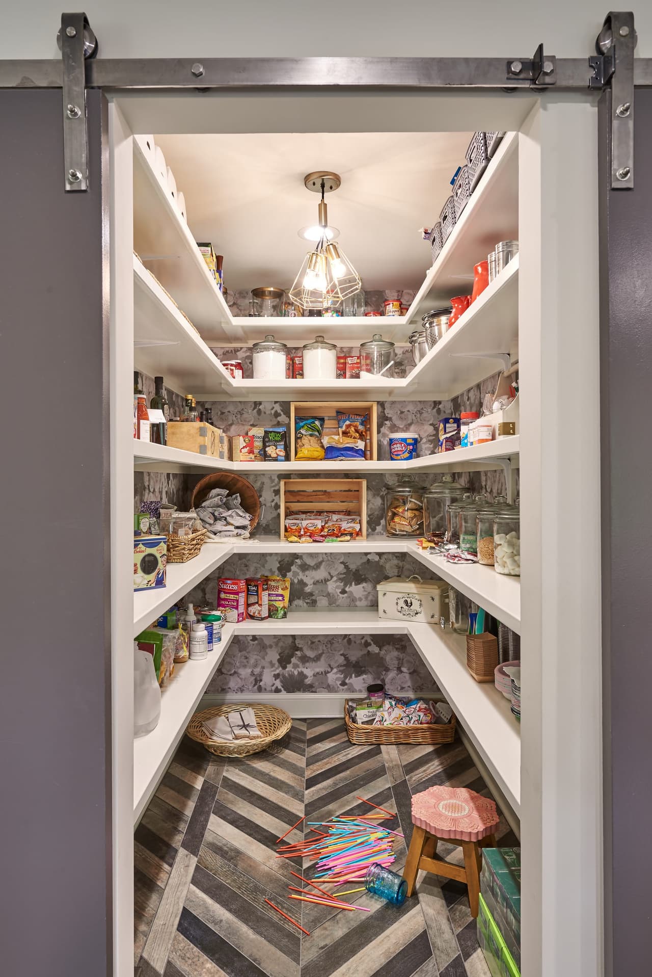 Walk-in pantry behind a barn door, with white shelving on three walls and a herringbone wood floor
