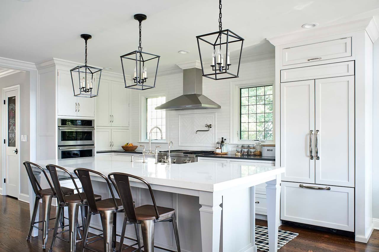 White kitchen in a Westfield, NJ modern farmhouse, with three iron lanterns over the island, counter stools and wall ovens beside the paneled refrigerator