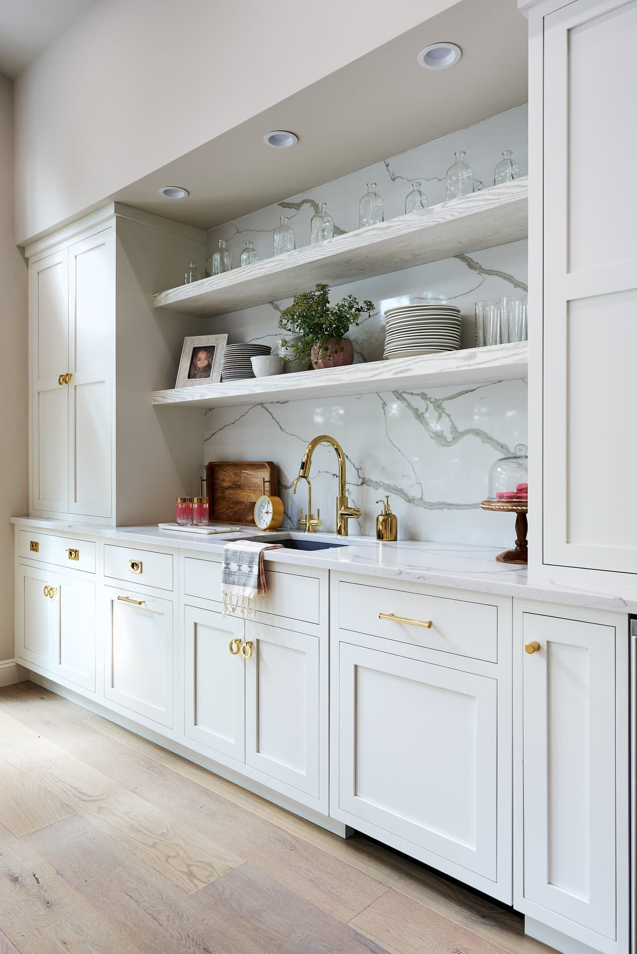 Butler's pantry with white cabinetry, open marble-backed shelving, a brass bar faucet and glassware on display