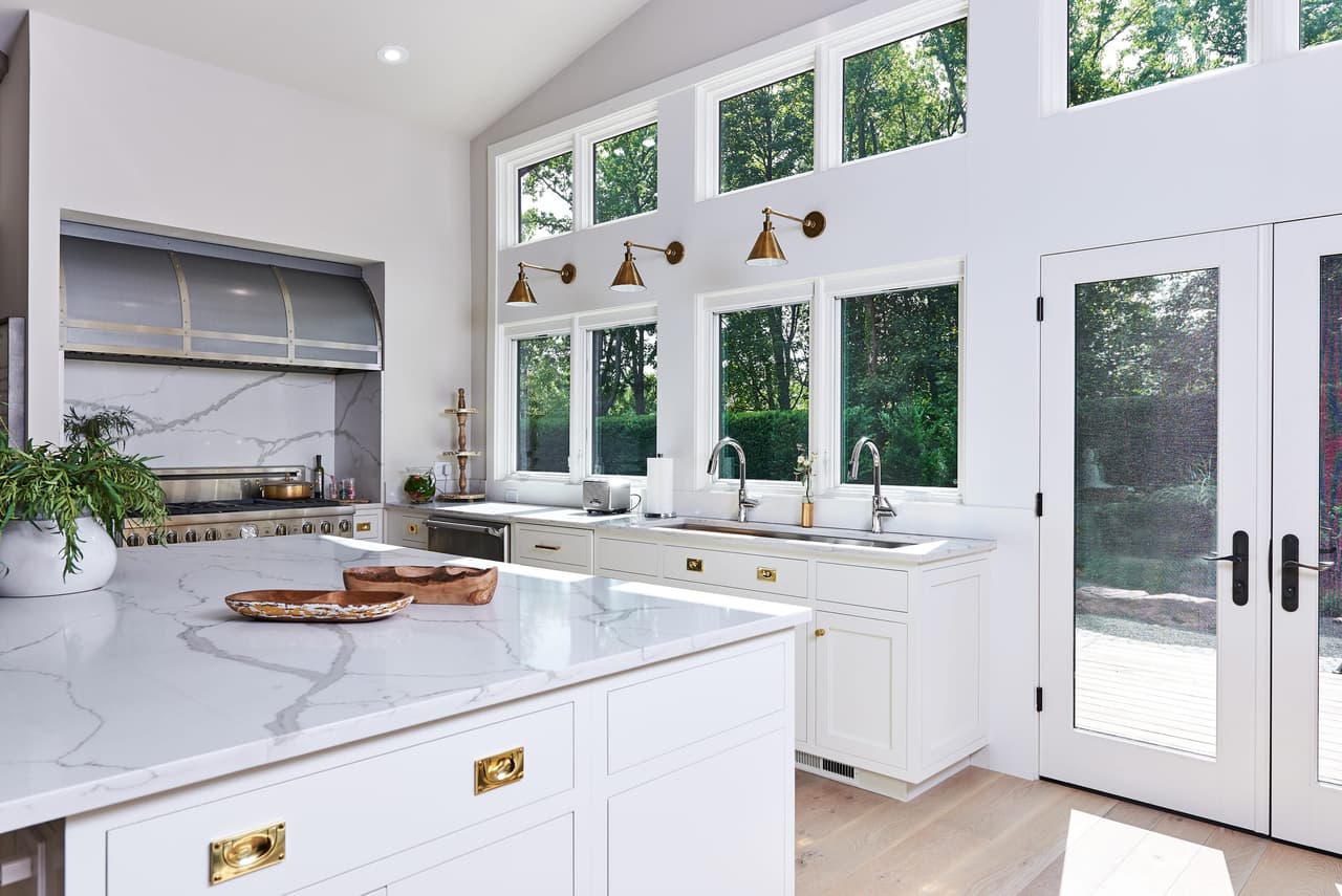 Kitchen with a wall of clerestory windows above the sink, a marble island and brass pulls on white cabinetry