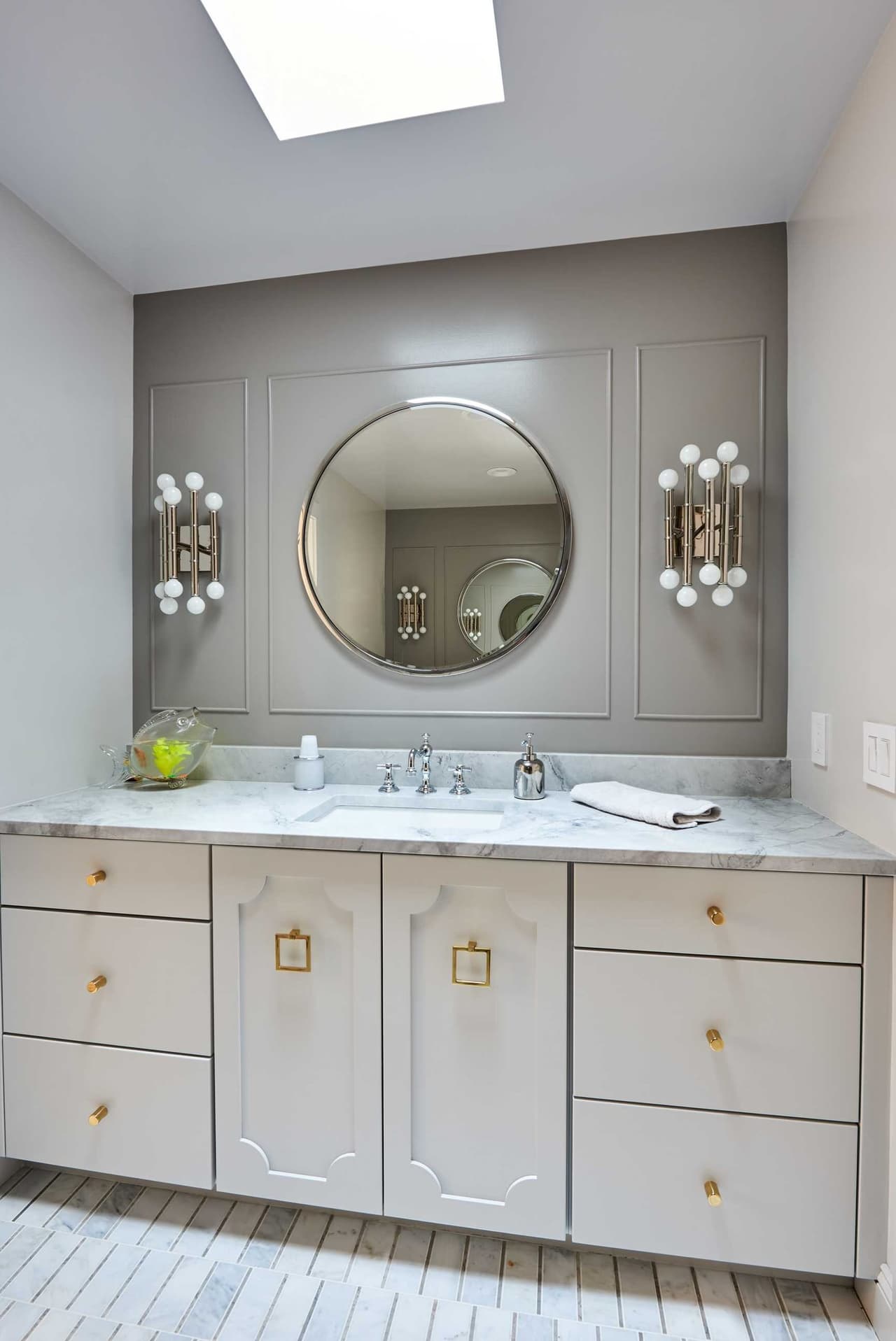 Bathroom with a round mirror between fluted sconces, a marble counter and gray cabinetry with brass hardware