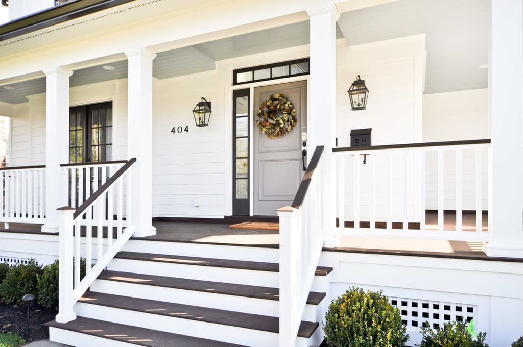 Farmhouse front porch with square white columns, lantern sconces and a gray front door under a transom