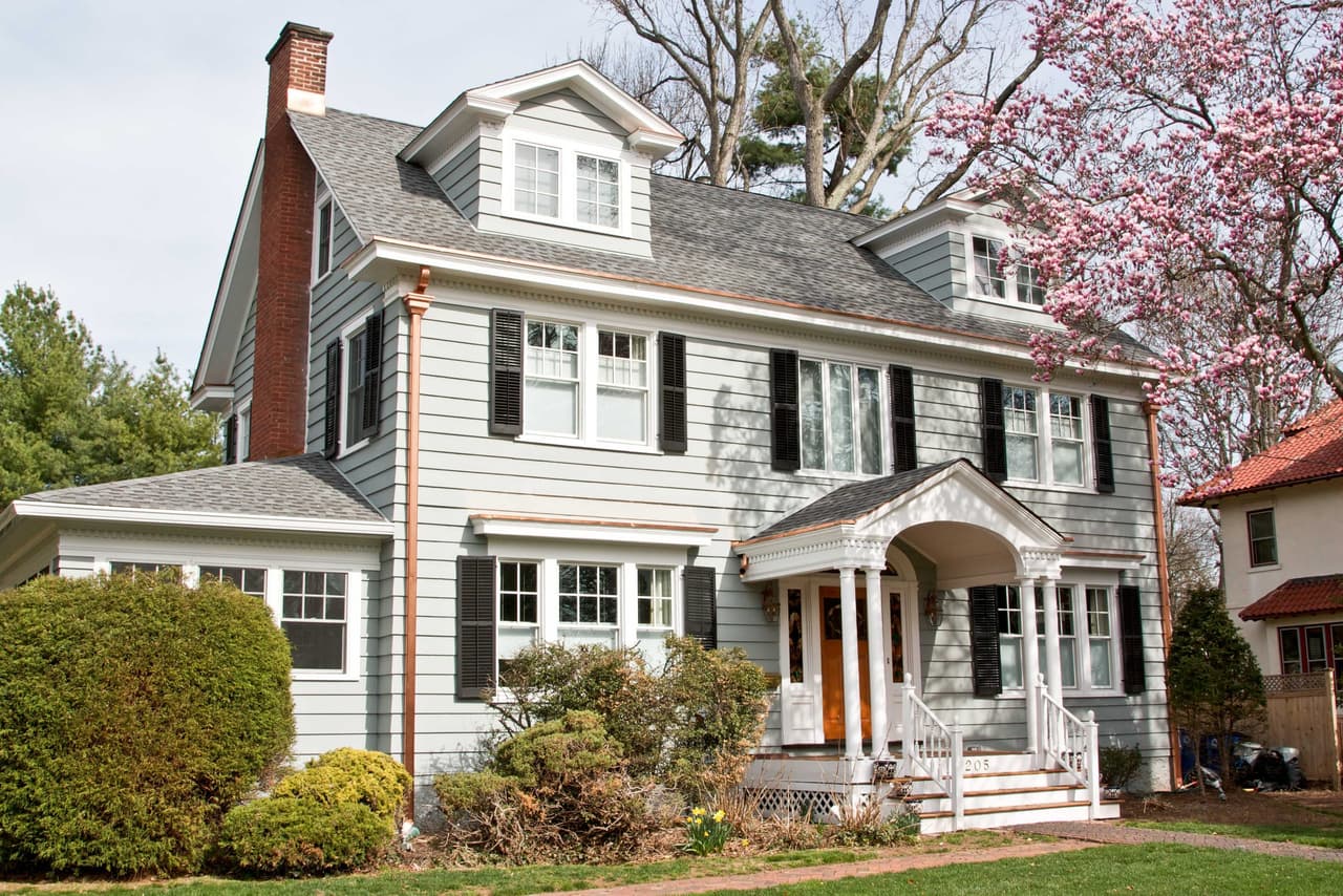 Front of a gray clapboard Westfield, NJ home with white trim, black shutters and two entry porches, with a cherry in blossom beside it