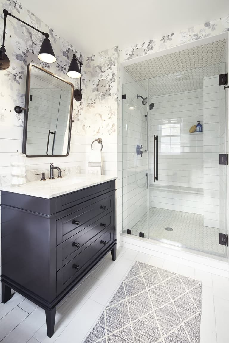 Bathroom with a dark vanity, a pivot mirror between black sconces, floral wallpaper and a glass shower