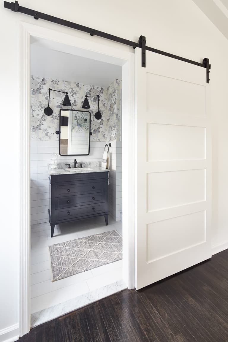 Bathroom seen through a white sliding barn door, with a dark vanity and patterned wallpaper