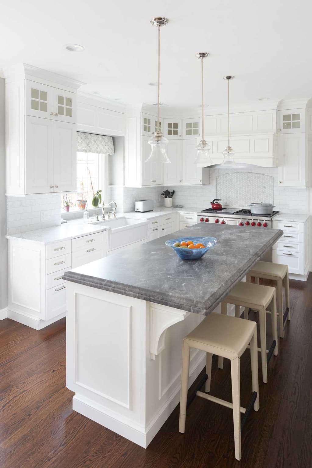 White kitchen in a Westfield, NJ renovation, with a soapstone island, three counter stools and glass-front upper cabinets either side of the window