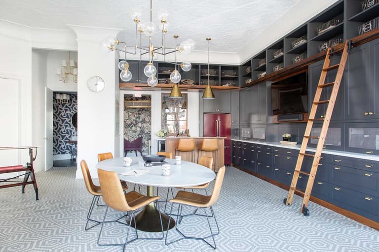 The MRCxEMD studio in Westfield, NJ, with a navy built-in cabinetry wall on a rolling library ladder, brass pendants and a round white table with tan leather chairs on patterned tile