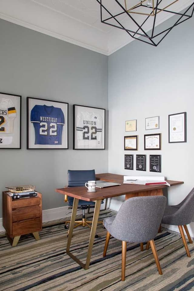 Office corner with a walnut desk, upholstered chairs, framed jerseys and a geometric pendant