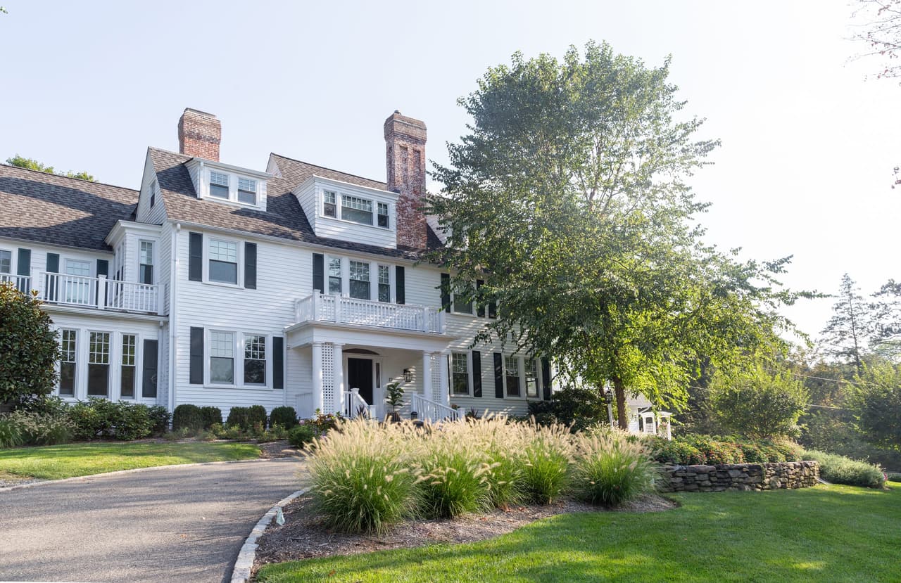 Front elevation of a white shingled historic home with twin brick chimneys, a columned porch and ornamental grasses along the drive in Short Hills, NJ