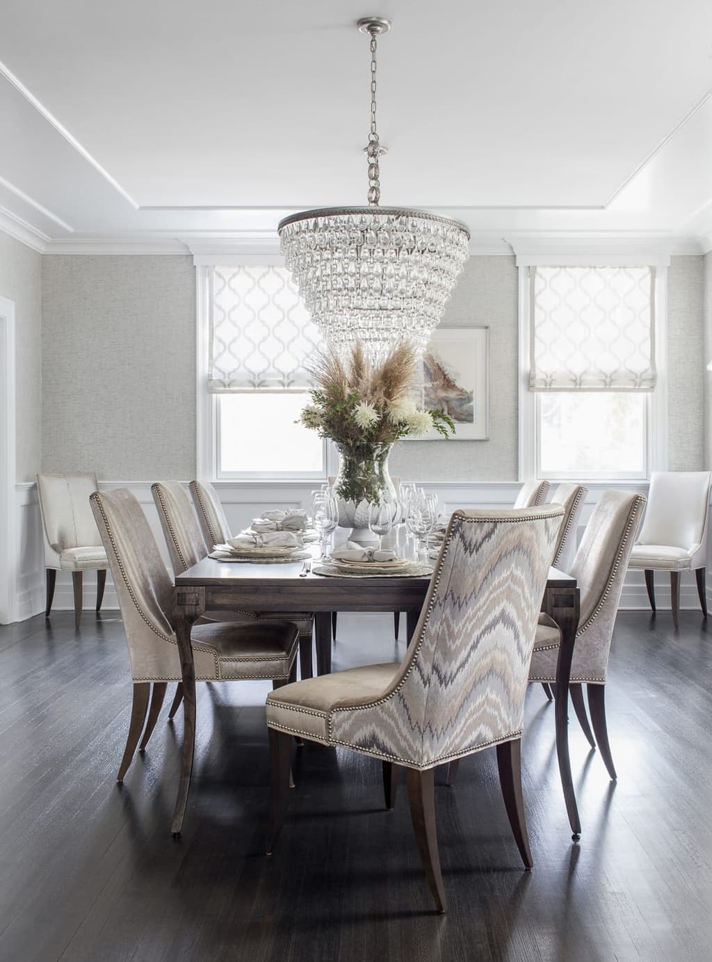 Dining room with a crystal drum chandelier, upholstered host chairs and woven Roman shades at the windows