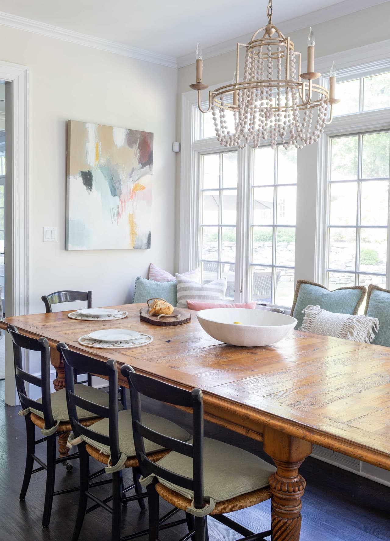 Breakfast room with a long farmhouse table, bentwood stools and a beaded chandelier beside a bank of windows with a built-in banquette