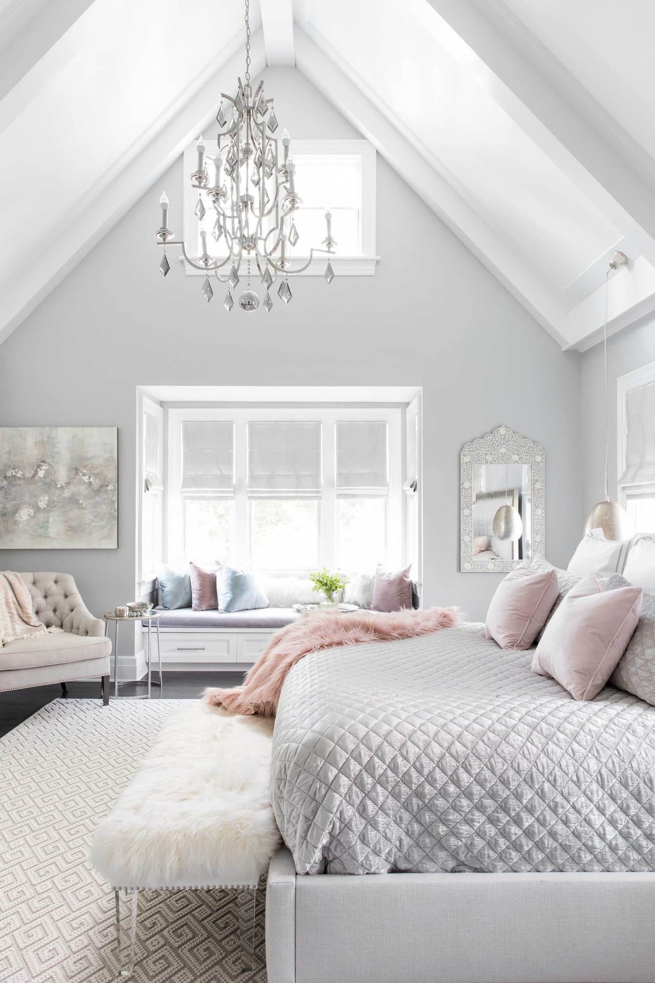 Bedroom under a vaulted ceiling with a crystal chandelier, a dormer window seat and an upholstered bench at the foot of the bed