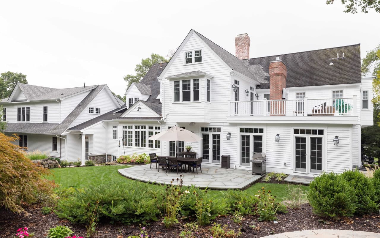 Rear elevation of the white shingled home with a stone terrace, gabled dormers and brick chimneys above the lawn in Short Hills, NJ