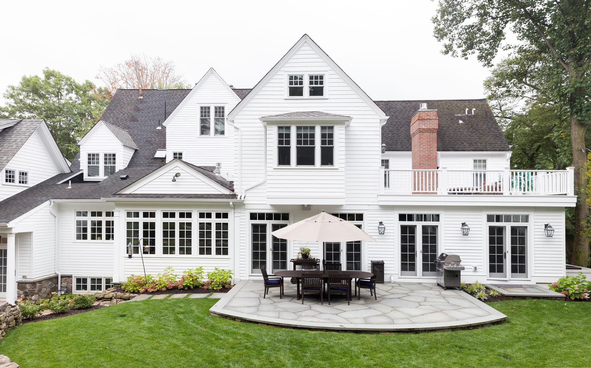 Rear of a white shingled home in Short Hills, NJ after renovation by Michael Robert Construction, with a brick chimney, a bluestone terrace and dining under an umbrella