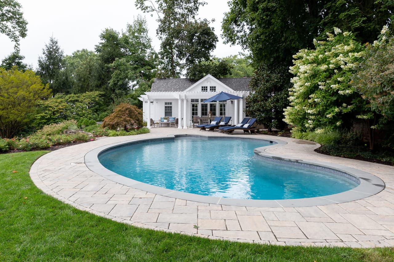 Kidney-shaped pool with paver coping set into the lawn, with the pool house and pergola beyond, in Short Hills, NJ