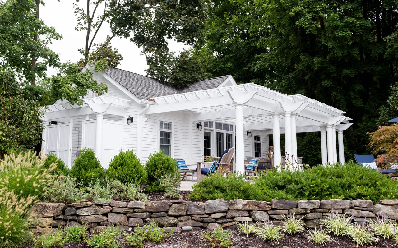 White pool house with a columned pergola above a dry-stacked stone retaining wall and planted border in Short Hills, NJ