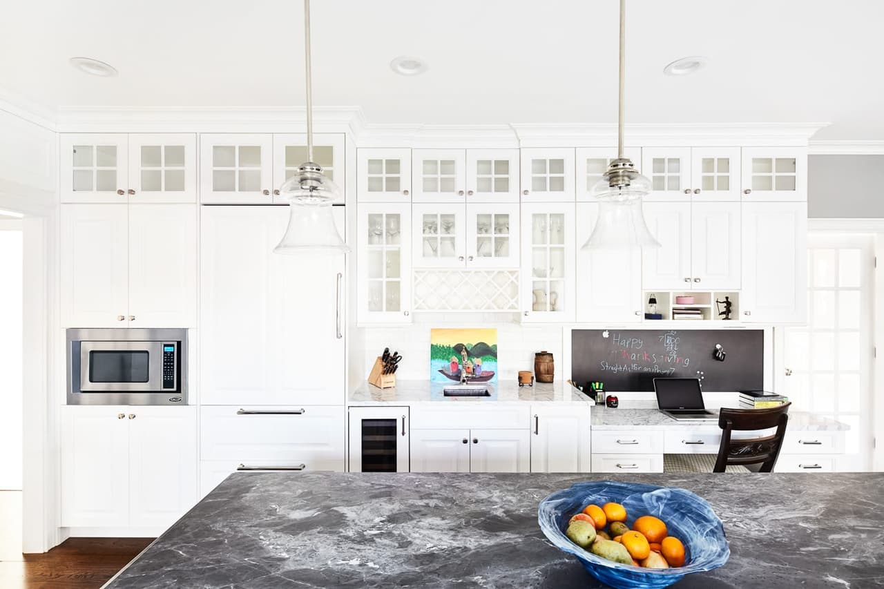 Kitchen with white cabinetry, glass-front uppers, a dark soapstone island and two glass pendants