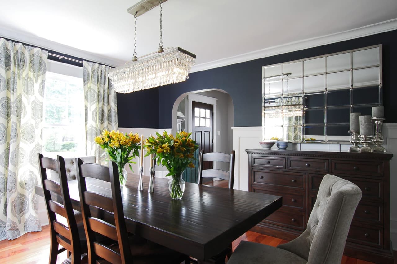 Dining room with a navy wall, a crystal linear chandelier and a dark sideboard beneath an arched opening