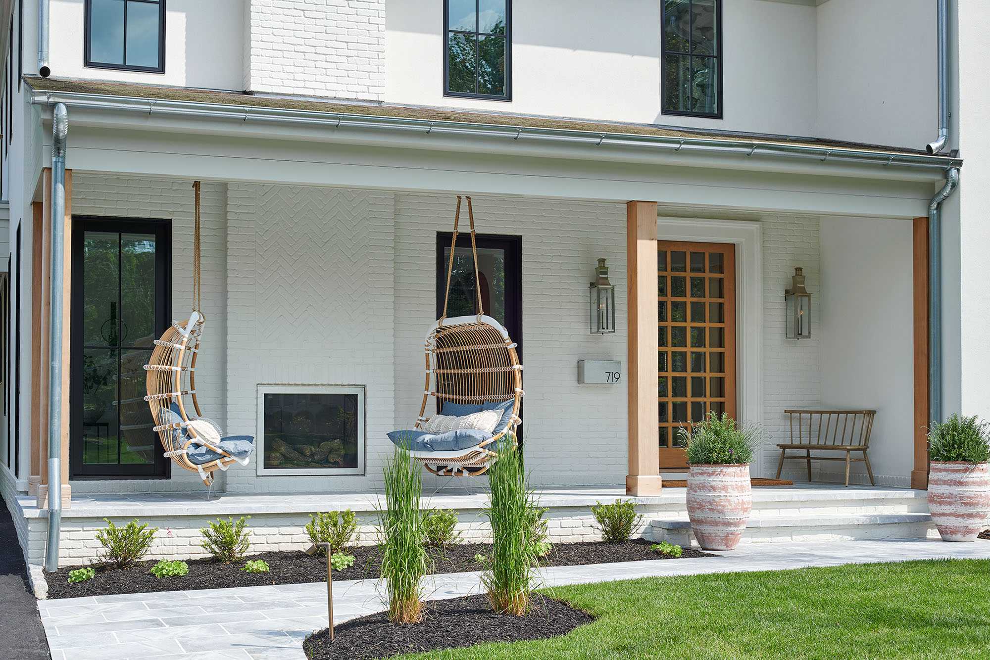 Front porch of a white painted brick home with cedar posts, hanging rattan chairs and a gridded wood front door