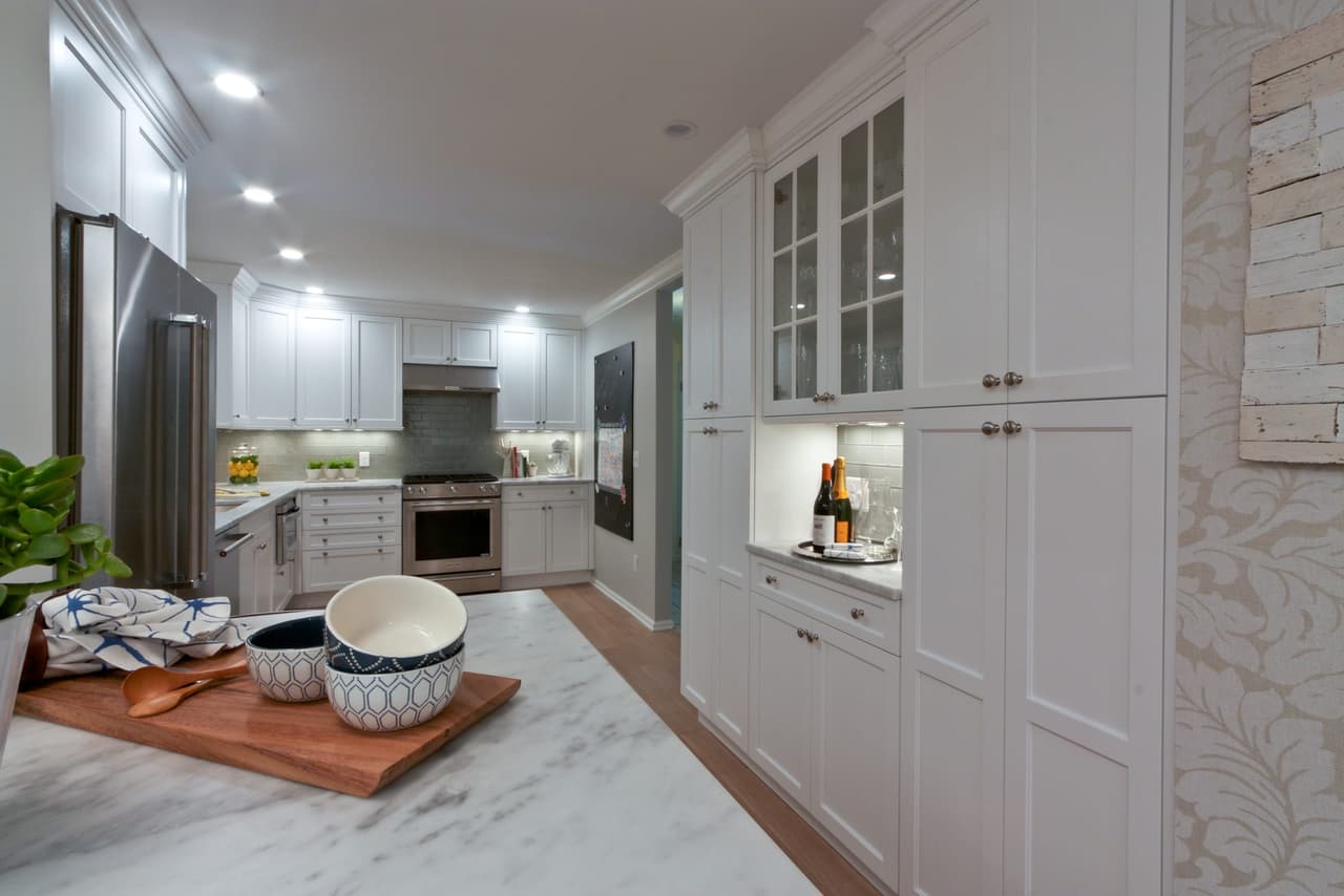 Marble counter with a wooden board and bowl, beside a run of white cabinetry and a glass-front cabinet