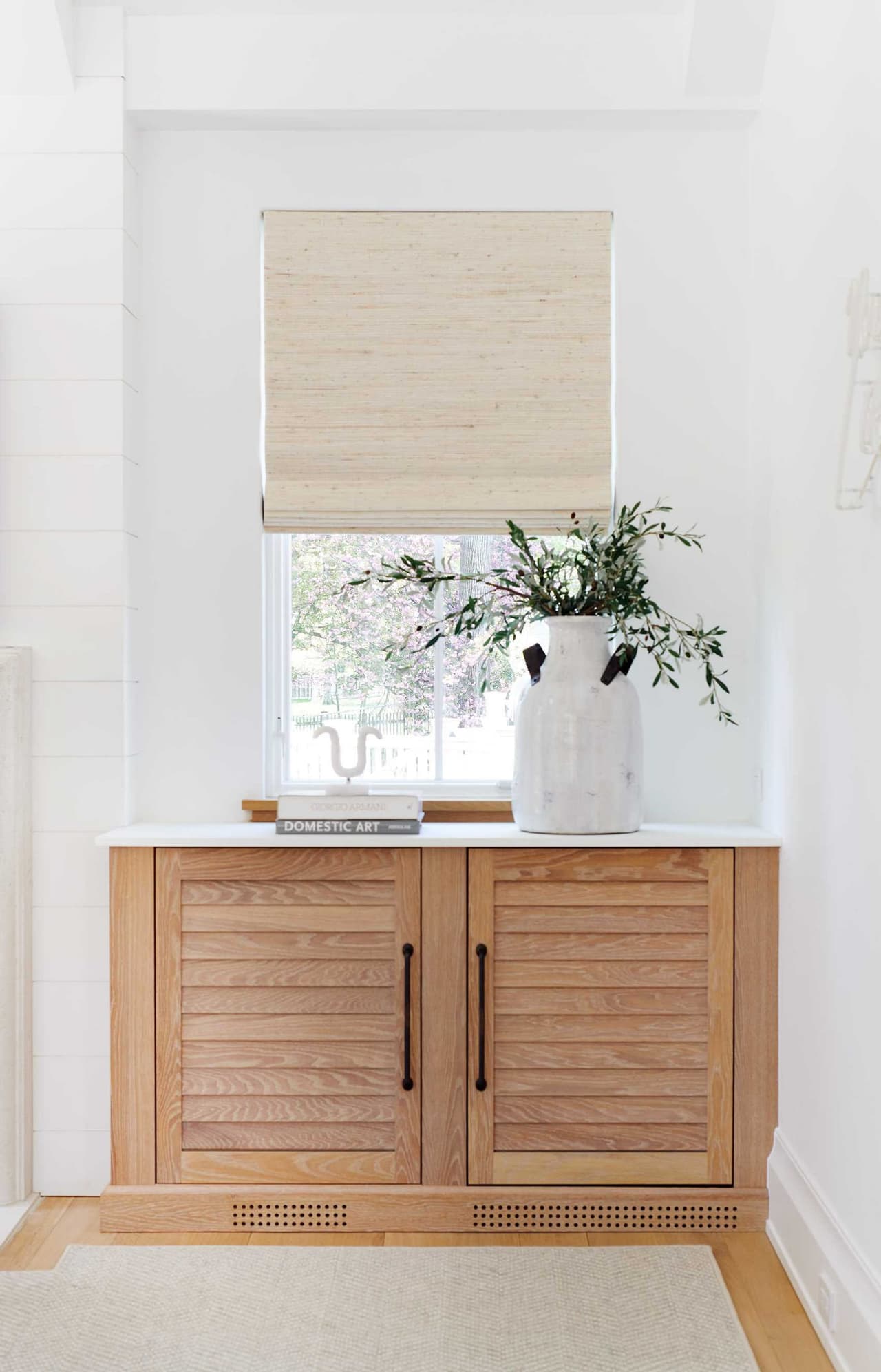 White oak louvered cabinet beneath a window with a woven shade, set against plaster walls