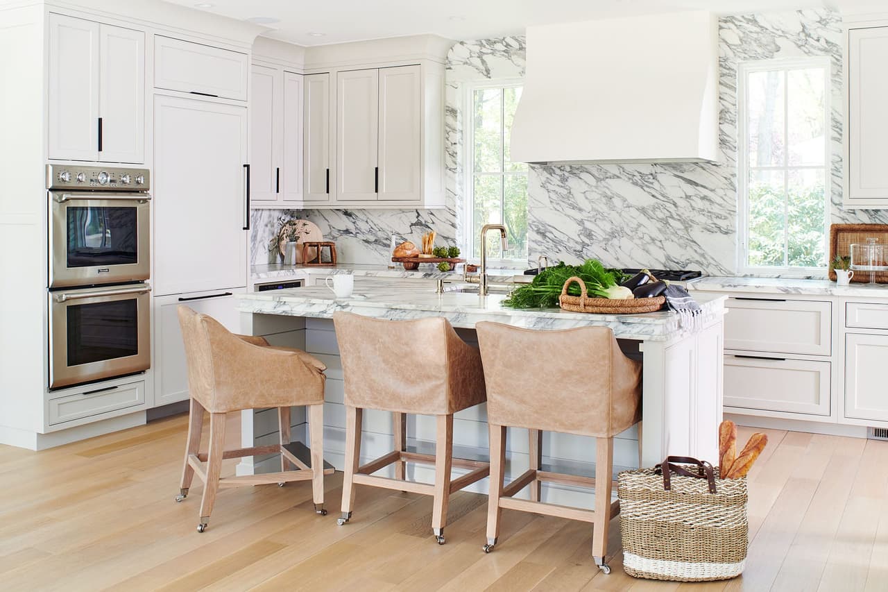 Kitchen of the Mroz family home in Westfield, NJ, with a marble island and leather counter stools, a plaster hood and wall ovens in the end run